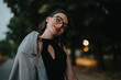 © qunica.com - Confident businesswoman with eyeglasses and gray jacket, smiling outdoors in the evening. Embracing professional yet relaxed style in a natural, pleasant environment.