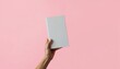 © umut hasanoglu - A hand holding up a blank white book against a pastel pink background, highlighting simplicity and minimalism.