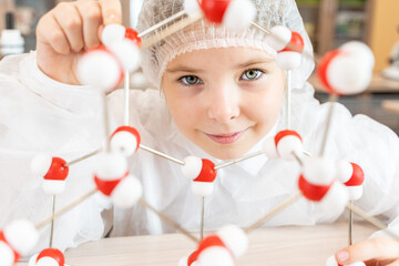 Portrait of a beautiful little schoolgirl in a white coat demonstrating an atomic model of a molecule using a set of three-dimensional atoms. Teenager studying chemical properties of elements in class