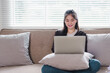 © kenchiro168 - Smiling young Asian woman uses laptop while sitting on couch, enjoying comfortable and relaxed moment at home.