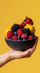 A hand presenting a black bowl filled with various fresh fruits and berries, including raspberries, blueberries, blackberries, and citrus against yellow.