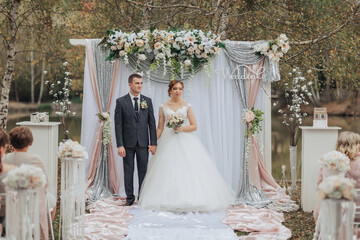 A bride and groom stand in front of a wedding arch. The bride is wearing a white dress and the groom is wearing a suit. The arch is decorated with flowers and has a silver color