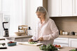 © fizkes - Happy mature woman spends weekend time in kitchen, cut fresh vegetables for vegetarian salad, try new dietary recipe, lead healthy lifestyle, enjoy culinary hobby, prepare delicious dinner for family