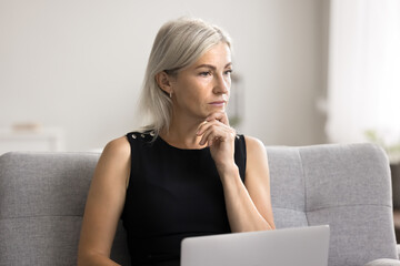 Puzzled mature woman sit on sofa with laptop, facing challenging task or problem looks deep concentrated, engaged in creative project, brainstorm idea or lack of understanding, search issue solution