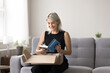 © fizkes - Smiling mature woman sitting on sofa unpacking box, get package, opening received parcel with ordered books using easy and comfortable on-line bookshop delivery services. Hobby, leisure, book lover