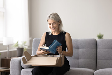 smiling mature woman sitting on sofa unpacking box, get package, opening received parcel with ordere