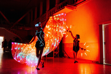 Two women are performing a light show with a large, glowing, multi-colored fabric. The scene is set in a large, brightly lit room with a red wall. The women are dressed in black