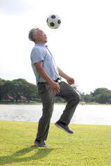 Happy elderly Asian man playing football in green public park. Joyful senior enjoys exercising outdoors with his favorite soccer sport. Active healthy elderly workout in outside garden nature backyard