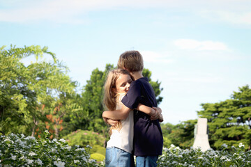 Happy family, brother hugging his little sister for showing love at beautiful flower garden. Joyful kids, boy and girl have fun outdoor at public park. Sibling children spending time outside together.