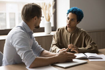 Attractive blue-haired African employee lead talk to male colleague seated at desk in office, looks serious while consider issue solution, exchanging opinions, ideas and creative thoughts at workplace