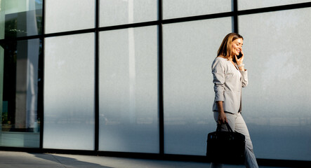 Wall Mural - Business woman strolls confidently outdoors in smart attire, checking her messages against the backdrop of a modern glass building on a sunny day