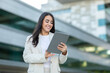 © Prostock-studio - A woman smiles while holding a tablet outside a contemporary building, showcasing a blend of technology and elegant fashion.