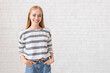 © Prostock-studio - Pretty blonde girl with hands in pockets smiling over white brick wall
