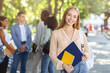 © Prostock-studio - Pretty smiling student girl with books posing at summer park, group of international chatting students on background