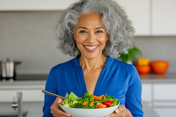 Happy elderly African woman with gray hair in blue blouse holding plate with a colorful vegetable salad in a modern kitchen. Healthy, plant-based lifestyle. Copy space.