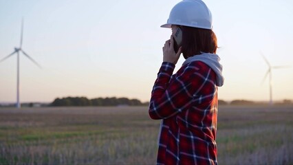 Female engineer talking on the phone in a field with wind turbines, as the sun sets. Concept of clean energy and engineering audit