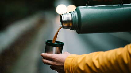 Close-up of man hand pouring coffee from green colored thermos into cup. Photo of metal stainless thermos in forest.