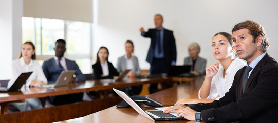 Portrait of serious focused middle-aged white brunette businessman sitting with young white female colleague beside at conference table in boardroom, intently watching business partner's presentation