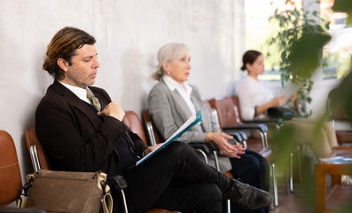 Wall Mural - Confident interested entrepreneur sitting in office hall waiting for meeting with investors or business partner, holding folder with documents..