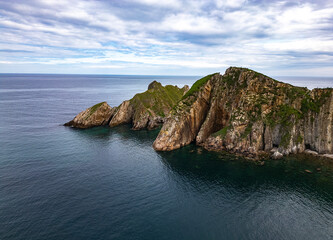 Aerial view of rugged coastal cliffs with green tops surrounded by calm blue waters under a cloudy sky