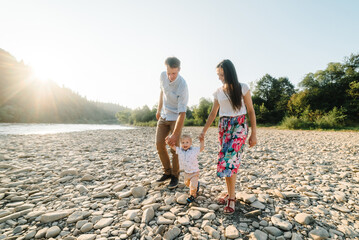 Mother, father hold hands baby son walking on stone beach near lake. Happy family holiday in summer. Father's, mother's, Children's day. Spending time together. Mom, dad embrace child at sunset.