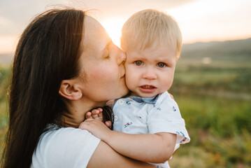 Mom embraces and kisses baby looking at the camera in sunset. Happy family holiday on a summer day. Mother hugs baby in the mountains. Parents hugging the child. Spend time together. Closeup face.