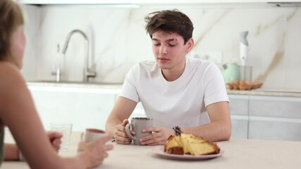 Wall Mural - Smiling young guy with her wife having breakfast and talking together at home kitchen