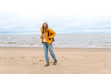 Wall Mural - Young woman in a yellow jacket smiles on the beach near the ocean, enjoying a windy day with gray skies. Calmness and tranquility. Travel, tourism concept.