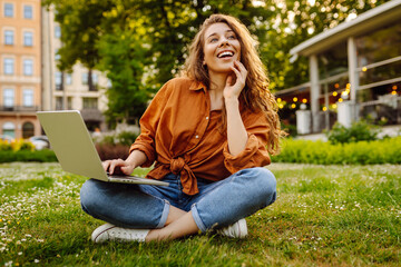 Wall Mural - A young woman enjoying a sunny afternoon working on her laptop in a park setting surrounded by greenery and urban views. Freelancer, Online education.