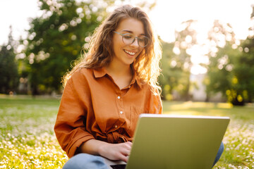 Wall Mural - A young woman enjoying a sunny afternoon working on her laptop in a park setting surrounded by greenery and urban views. Freelancer, Online education.