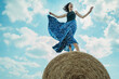 © Westend61 - Carefree mature woman wearing skirt standing on hay bale under cloudy sky