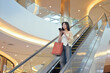 © Prathankarnpap - Pretty young asian woman with shopping bags standing on escalator and using mobile phone