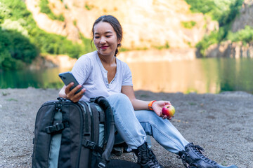 a young woman sat down to rest by the shore of a lake after a hard hike checking her phone, having a snack next to her equipment