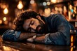 © DKPhoto - A weary man in a denim shirt rests his head on the bar counter of a dimly lit bar. The atmosphere suggests quiet contemplation or a long day ending in solitude.