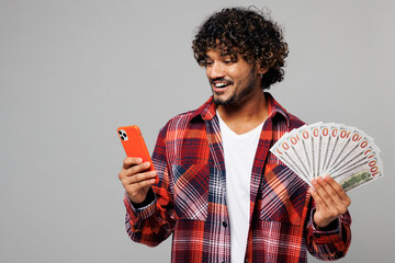 Sticker - Young happy smiling Indian man he wearing red shirt casual clothes use mobile cell phone hold in hand fan of cash money in dollar banknotes isolated on plain grey color background. Lifestyle concept.