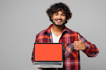 Young Indian IT man wears red shirt casual clothes hold use work on laptop pc computer with blank screen workspace area show thumb up isolated on plain grey color background studio. Lifestyle concept.