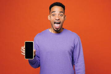 Poster - Young happy amazed man of African American ethnicity wear purple sweatshirt casual clothes hold in hand use mobile cell phone isolated on plain red orange background studio portrait Lifestyle concept