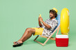 © ViDi Studio - Full body side view young happy Indian man wear beach shirt hat rest near hotel pool sit in deckchair hold use mobile cell phone isolated on plain green color background. Summer sea sun tan concept.