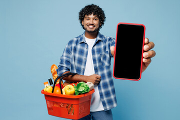 Wall Mural - Young Indian man he wear shirt hold red basket with food products vegetables use blank screen area mobile cell phone isolated on plain pastel blue background. Delivery service from shop or restaurant.