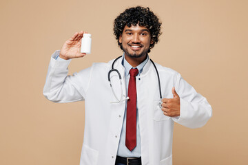 Young Indian doctor man wear white medical gown suit work in hospital clinic office hold jar of drugs medication show thumb up isolated on plain beige background studio. Healthcare medicine concept.