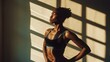 © VK Studio - A woman in a sports bra poses confidently in sunlight streaming through blinds, emphasizing her defined muscles and focused determination.