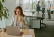 © Yaroslav Astakhov - A young woman enthusiastically gives a thumbs up while diligently working on her laptop in a bright office environment