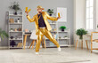 © Studio Romantic - Excited overjoyed funny young man holding gold bar having fun in living room at home celebrating success or win a lottery. Rich guy wearing golden suit dancing indoors. Investment concept.
