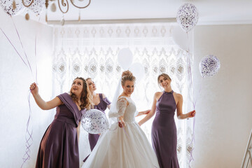 A bride and her bridesmaids are playing with balloons in a room. The bride is wearing a white dress and the bridesmaids are wearing purple dresses. Scene is lighthearted and fun