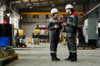 © TrueFrame Collective - Two industrial workers in safety gear discussing work tasks inside large warehouse with machinery and equipment