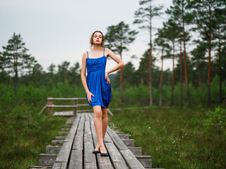 Young teenager girl in high fashion blue dress posing in a forest park on a wooden footpath, selective focus. Prom photo shoot. Model short hair and slim body type.