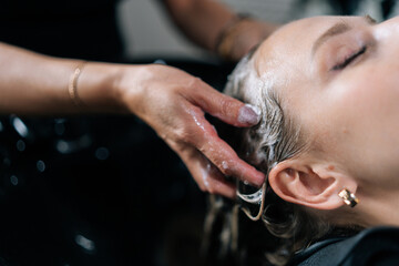 Wall Mural - Closeup hands of hairdresser washing head of young woman in beauty hairdressing salon. Close-up of unrecognizable hairstylist foaming shampoo on long blonde hair of beautiful female client.