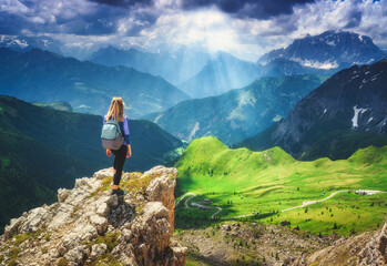 Wall Mural - Girl with backpack on the rock in beautiful alpine mountain valley, green meadows at sunset in summer. Landscape with young woman in alps, dramatic sky with clouds, sun rays. Dolomites, Italy. Hiking