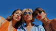 © THINGDSGN - Three stylish friends pose together against a blue sky on a sunny day, warmth and joy of friendship, trendy summer fashion and stylish sunglasses