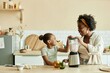 © Seventyfour - Side view of smiling African American mom and little daughter having fun while cooking breakfast together making smoothie in blender on large wooden counter at cozy beige kitchen, copy space